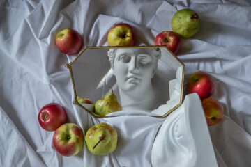 Ancient greek sculpture white plaster marble head of diana and eye of david reflected in a mirror in a frame against a background of white drapery fabric with folds with red apples and green pears