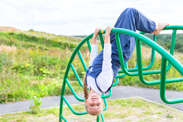 Cheerful girl in a Blue summer overalls hanging upside down on Green playground, looking at camera, smiling.