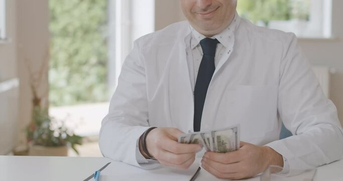 Unrecognizable happy doctor counting dollars and smiling. Satisfied Caucasian male physician calculating earnings or bribes in office. Wealthy man working in medicine. Cinema 4k ProRes HQ.