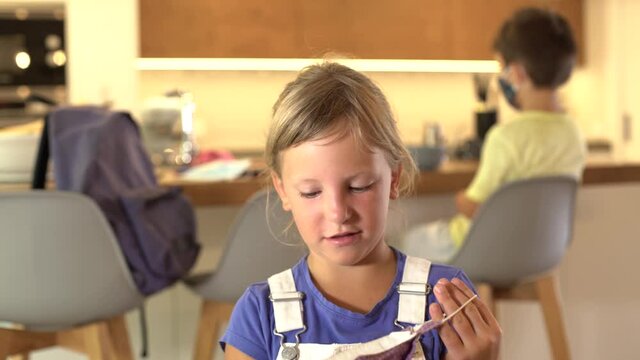 Caucasian female kid puts on a protective mask at home looking at camera on First day School at the kitchen at home. A little boy behind has his protective mask on. Concept new rules Coronavirus.