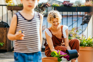 Amazing blond positive woman gardener at her balcony during planting. Her son running on foreground. People, gardening, flower planting and profession concept.
