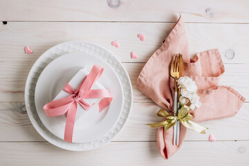Festive table setting. Plates and cutlery with pink napkin and gift box on white wooden background.