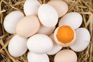 Chicken eggs of white and brown color with a broken egg in the center, in which a fresh yolk is visible, lie on the straw. The view from the top. Food background. Concept of natural farm products.