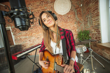 Fototapeta na wymiar Cheerful. Beautiful woman in headphones recording music, singing and playing guitar while sitting in loft workplace or at home. Concept of hobby, music, art and creation. Creating first single.