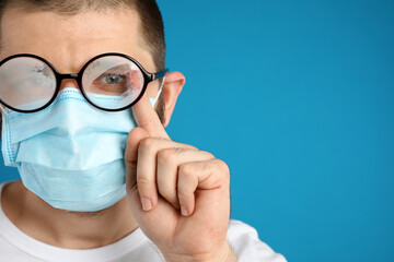 Man wiping foggy glasses caused by wearing disposable mask on blue background, space for text....