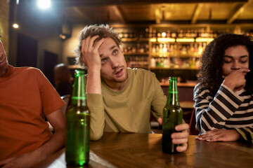 Young man looking disappointed while watching sports match on TV, drinking beer and cheering for team in the bar. People, leisure and entertainment concept