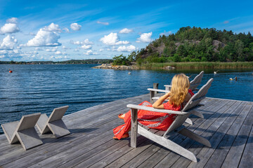 A blonde woman in a red long dress on the seaside bay. A beautiful girl is resting outdoors sitting in a chair on a wooden pier. The female in red relaxing and looking at the water. Summer vacation.