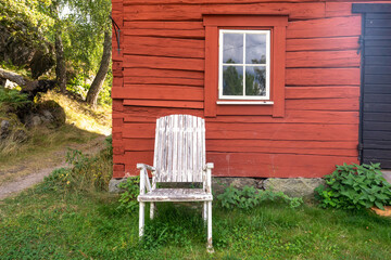 Fototapeta na wymiar Old Swedish red wooden house in a village. Aged shabby wooden outdoor chair of white color for recreation. Traditional rural or countryside summer homes in Scandinavia. Landscape garden design.