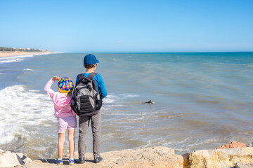 Back view of brother and sister playing on the beach holding hands together. Beautiful day time activity, sunny outdoors background.