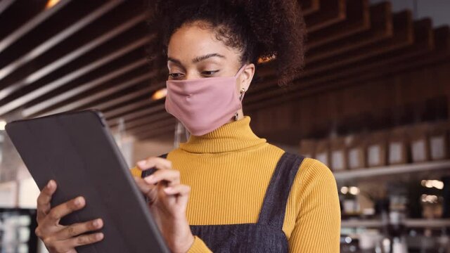 Medium Close Up, Female African coffee shop owner wearing face mask standing in front of counter using digital tablet for stock check..