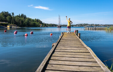 Obraz premium Young woman stands on a wooden pier on the sea bay and waves to moving sailing yacht. Slender beautiful Girl in shorts. Blond Female on shore in the Baltic sea bay in Scandinavia. Panorama.