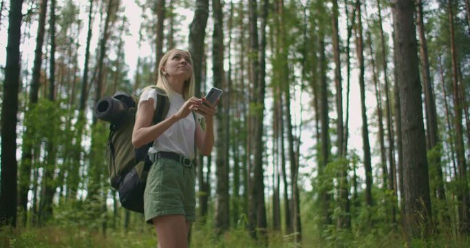 A young woman with a mobile phone walks through the forest traveling with a backpack in slow motion. Navigate through the forest using the Navigator in your mobile phone