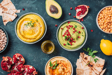 Bowls with yellow hummus and green hummus, tahini, olive oil, sesame seeds, pita, raw chickpeas, avocado, pomegranate on dark background. Middle eastern, jewish, arabic cuisine. Top view