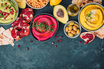 Ingredients for yellow turmeric hummus, green parsley hummus and red beetroot hummus on dark background, lemon, sesame, olive oil, pita, avocado, pomegranate. Middle eastern, jewish, arabic cuisine.