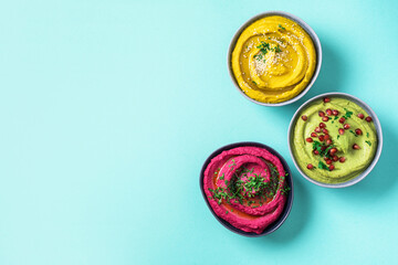 Colorful hummus bowls - green, yellow and beetroot hummus on dark background with lemon, olive oil, sesame seeds, pita, raw chickpeas, avocado, pomegranate. Middle eastern, jewish, arabic cuisine.