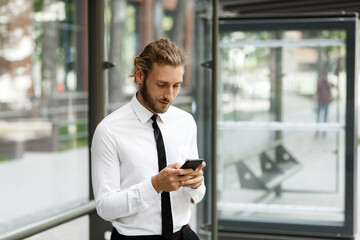 A curly-haired guy, in a white shirt and tie, at the bus stop looks at the phone while waiting for the bus. The concept of a mobile application for traffic in the city.