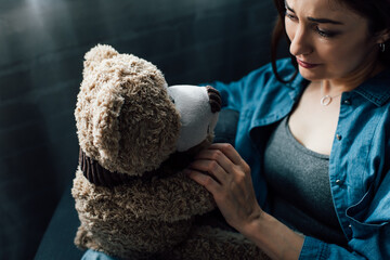 upset brunette woman holding teddy bear and crying at home