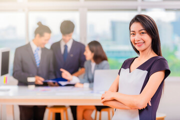 Beautiful young asian woman in office folded arms and confident expression as other workers hold a meeting in background.