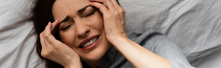 panoramic shot of brunette woman suffering from pain and touching head while lying on bed