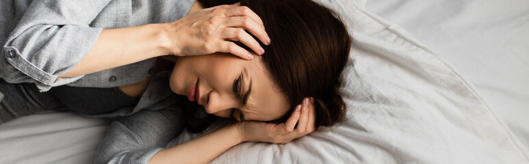 panoramic shot of brunette woman having migraine, touching head and lying on bed