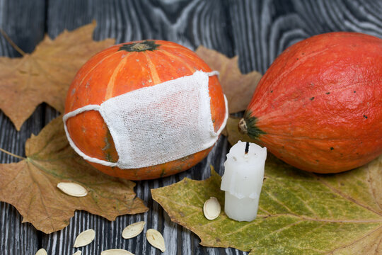 Orange pumpkin with a medical mask. Nearby is another pumpkin and a candle. Pumpkin seeds and dried maple leaves are scattered. Against the background of brushed pine boards painted in black and white