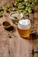 Classic glass mug of fresh cold foamy lager beer with green hop cones, wheat grain and red fermented malt in ceramic bowls behind over wooden texture background. Copy space