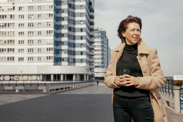 Fototapeta premium Woman in beige coat walks along the road along modern high-rise buildings