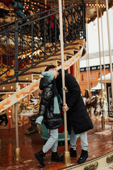 Young couple in warm clothes embracing and kissing outdoors on the carousel  in amusement park....