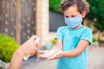 Mother and kid with medical mask disinfecting hands with sanitizer outdoors. Life during coronavirus pandemic.