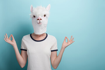 Young woman wearing alpaca mask. Hands in meditation yoga mudra sign. Isolated on blue background.