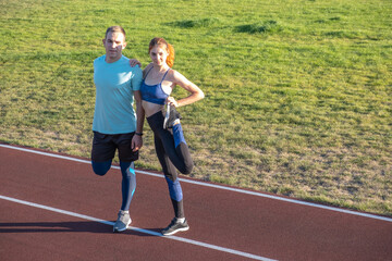 Young couple of fit sportsmen boy and girl doing warming up exercises  before running on red tracks of public stadium outdoors.