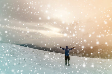 Back view of tourist hiker in warm clothing with backpack standing with raised arms on clearing covered with snow on spruce forest mountain and cloudy sky copy space background.