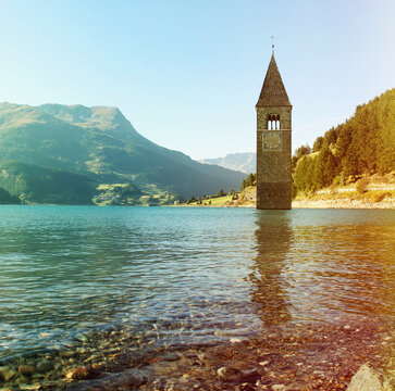 Famous sunken bell tower in Reschensee lake, South Tyrol, Italy. Submerged church spire in Lago di Resia water with alpine mountain landscape backdrop, famous travel landmark.