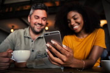 Cute smiling mixed race couple looking at smartphone while holding a warm cup of coffee sitting at trendy coffee shop.