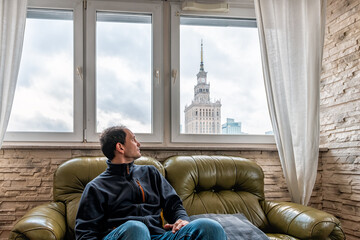 Young man sitting on couch in flat apartment looking at view of Warsaw, Poland famous Palace of...