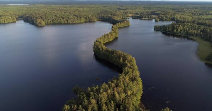 Aerial view overlooking a long, narrow, green Kyynaranharju esker at a lake, on a sunny, summer evening, in Hame, Finland