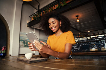Fototapeta na wymiar Low angle shot of beautiful African American female holding digital tablet sitting at funky tearoom with hot latte.