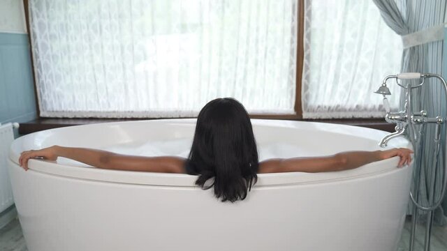 Back view of African woman lying in bath and looking at window. Spa and relaxation concept. Dolly out shot. Camera moving backward from woman