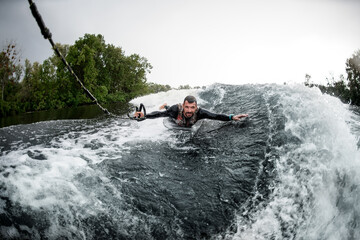 Obraz premium man holding rope and lying on wake surfboard rides on wave from motor boat