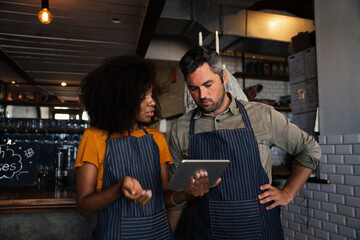Curious colleagues wearing aprons, discussing finances while looking at tablet standing in coffee shop. 