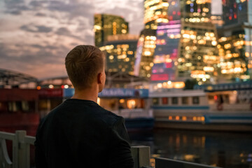 A business man in a black sweater stands with his back to the camera with a view of the night illumination business skyscrapers in Moscow