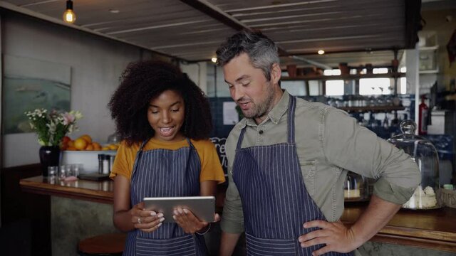 Good looking cafe owners, wearing aprons, discussing projects on tablet while standing in trendy coffee shop.