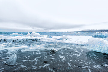 Waves shore long exposure motion on blue glacier iceberg ice floating on black sand by Jokulsaron lagoon lake diamond beach in Iceland