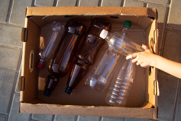 The child's hand puts a white bottle in the box. Plastic bottles are collected in a cardboard box for further disposal. Taking care of environment.