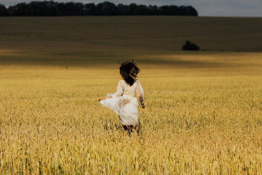 A beautiful cheerful girl runs along a wheat field in a beige dress. Back view. 