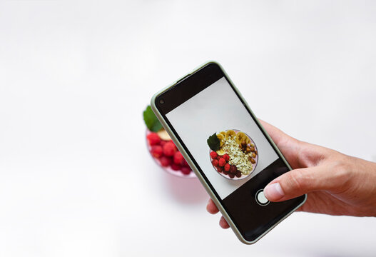 The blogger's male hand takes pictures of raspberry and fresh raspberry smoothies, banana, yogurt, nuts, oatmeal on a smartphone. Vegetarian concept, healthy food and diet. Top view, copy space. 