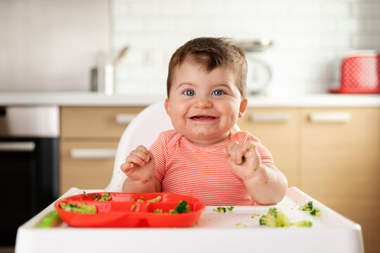 Funny smiling baby making mess while eating broccoli with hands