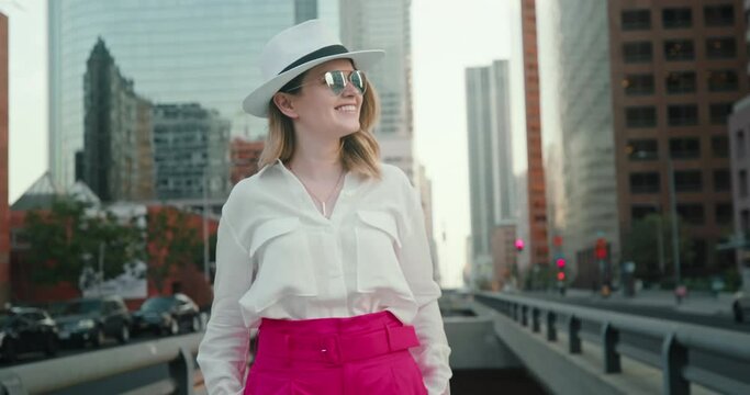 Smiling female traveler exploring Los Angeles downtown. Pretty stylish girl with urban view on background. Woman is sightseeing skyscrapers in California business district, 4K slow motion. USA travel