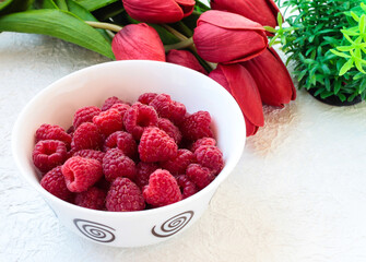 Raspberries in a white bowl on a light background with tulips.
