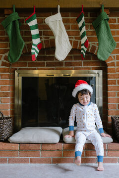 boy in pajamas and Santa hat sits in front of fireplace with stockings